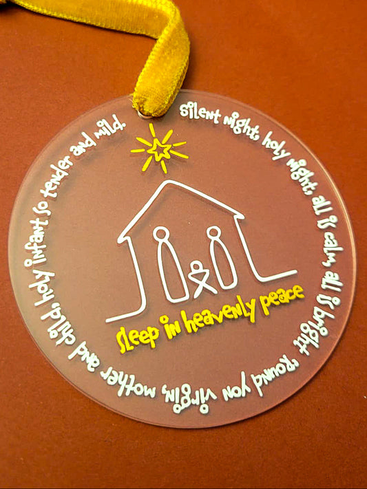 3 inch round ornament depicting a white minimalistic nativity scene with Joseph, Mary and Baby Joseph. I white circling the ornament is the lyrics to Silent Night. Under the nativity scene in gold, it says "Sleep in Heavenly Peace" and a gold star above the figures. The Ornament has a gold velvet ribbon at the top. 